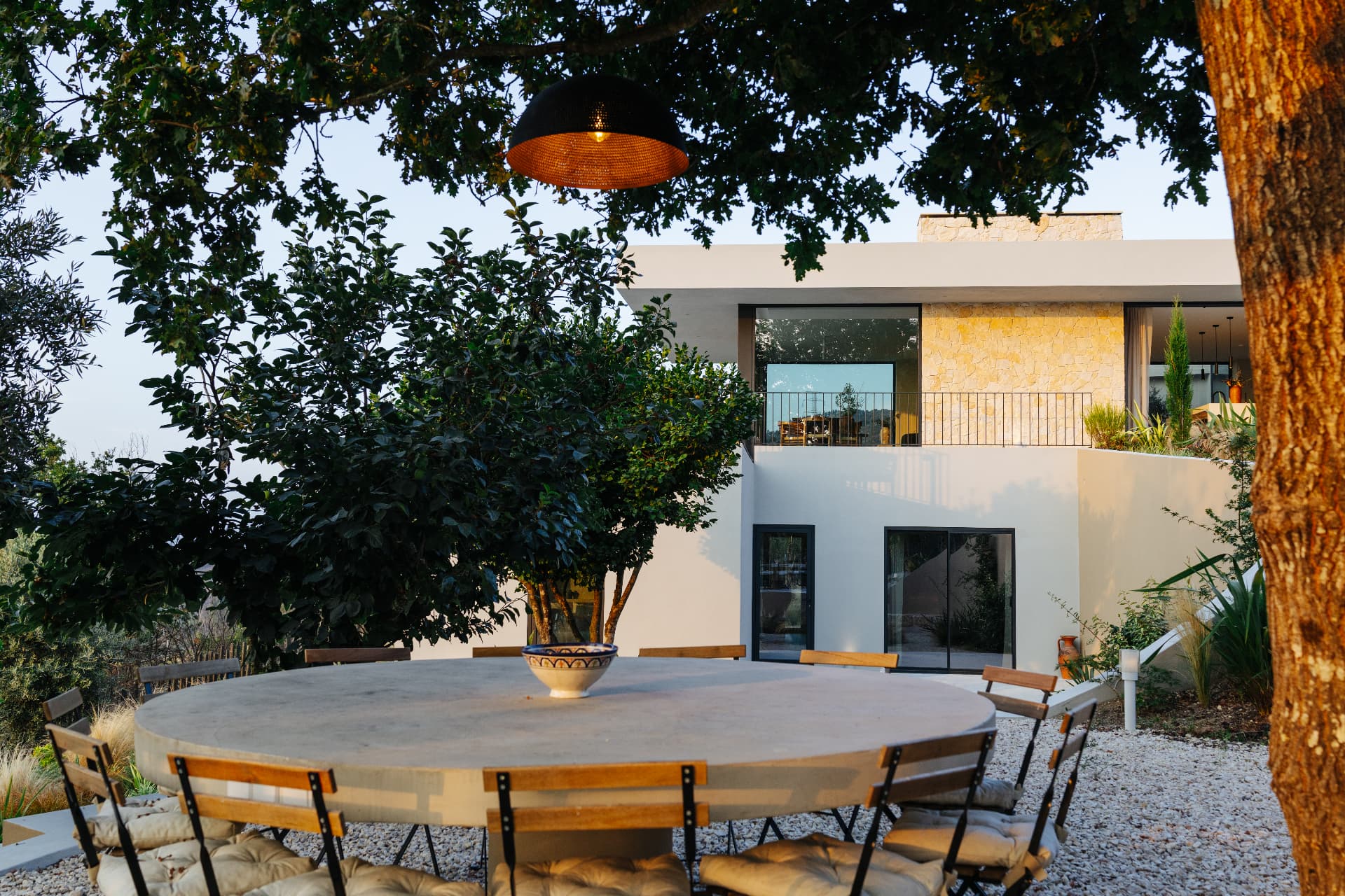 Round outdoor dining table beneath a tree with the villa and garden behind